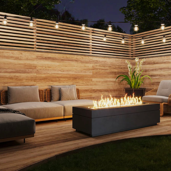 Boxhill's Linea fire pit burns bright as the centerpiece on a backyard deck with wooden privacy wall and string lights.