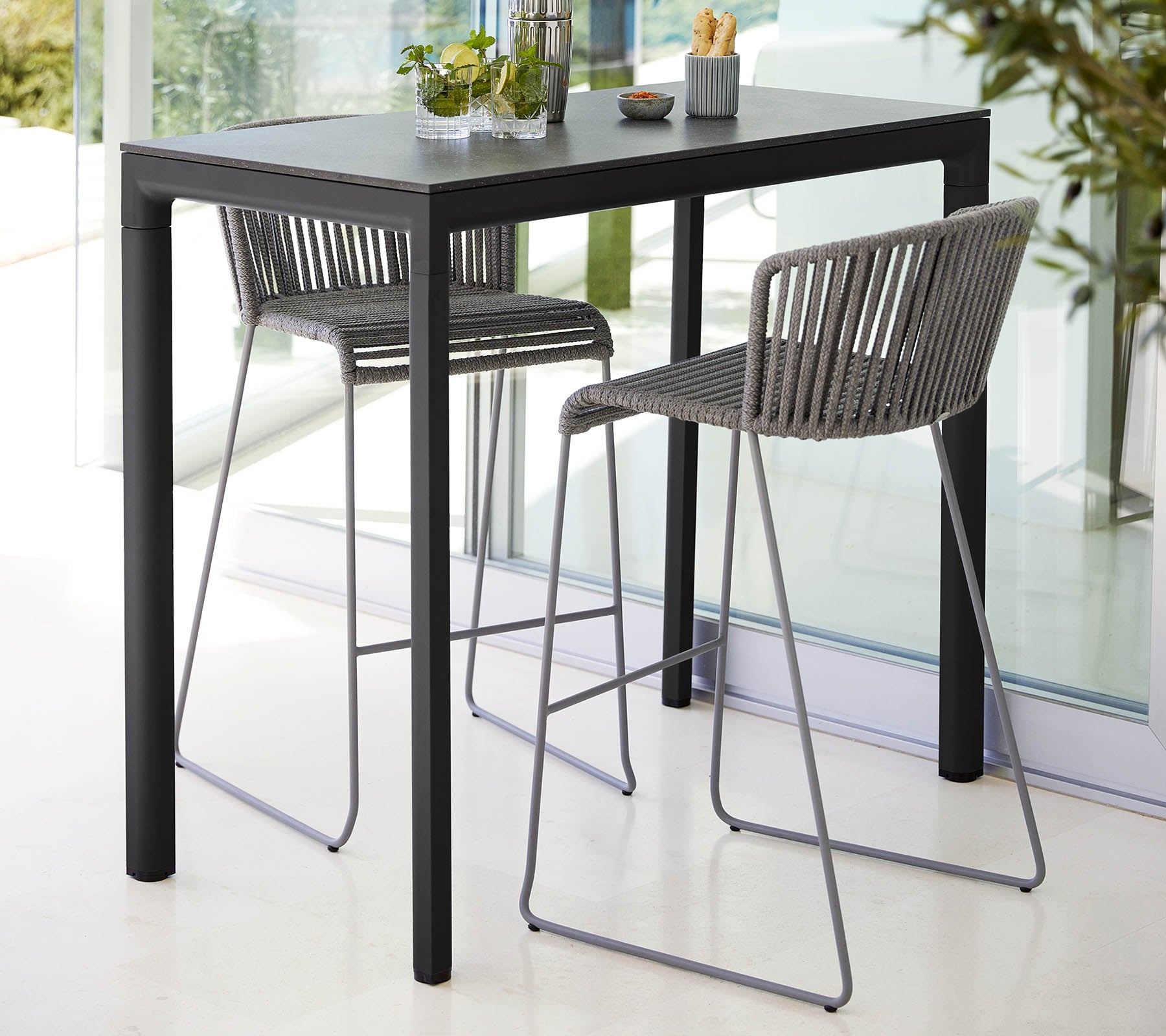 Boxhill's Drop Outdoor Bar Table Lava Grey lifestyle image with Breeze Bar Stackable Chair beside glass wall