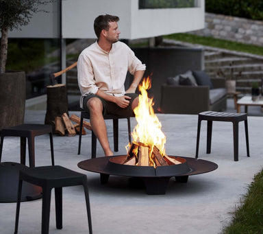 Boxhill's Cut High Outdoor Stool lifestyle image with a man sitting down and a fire pit in front. 