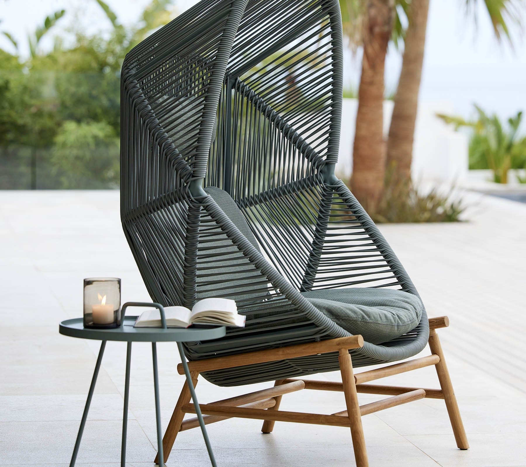 oxhill's Hive Outdoor Hanging Chair Dusty Green Frame lifestyle image with round side table at the side with candle inside the glass and a book on top