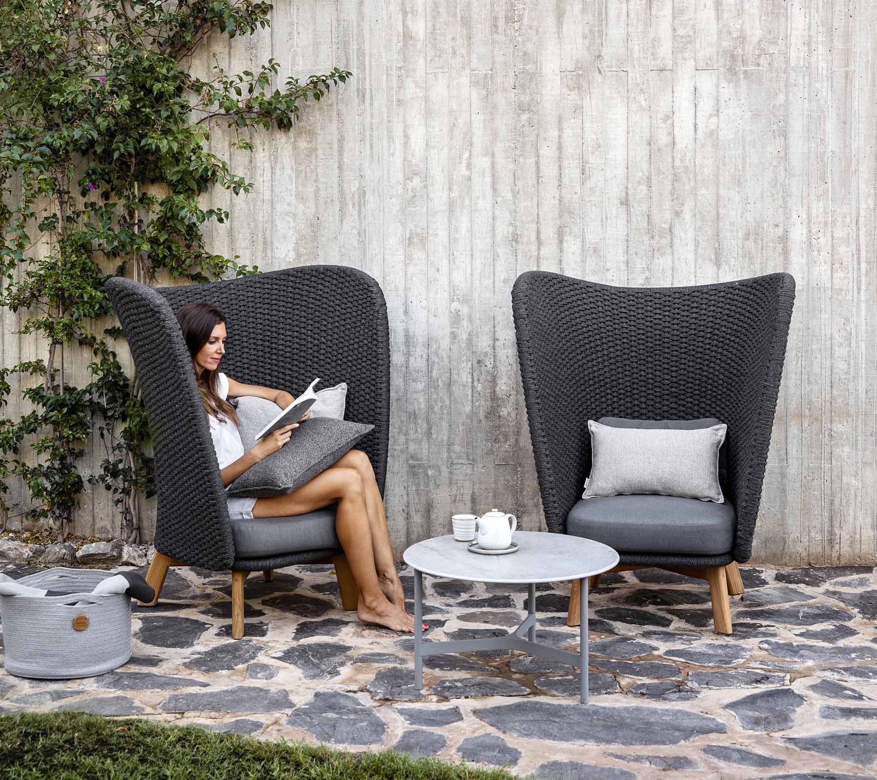  Boxhill's Peacock dark grey outdoor wing highback chair with a woman sitting on it reading a book and light grey coffee table