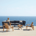 Boxhill's Conic 2-Seater Right Module Sofa Grey lifestyle image on wooden platform with a woman sitting down at seafront