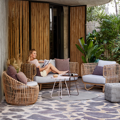 Boxhill's Nest 2-Seater Sofa lifestyle image at patio with Nest Lounge Chair, Nest Round Rattan Chair and a woman sitting down reading a magazine