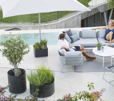 Boxhill's lava grey outdoor round large modern planter box with man and woman sitting on light grey sectional sofa beside the pool