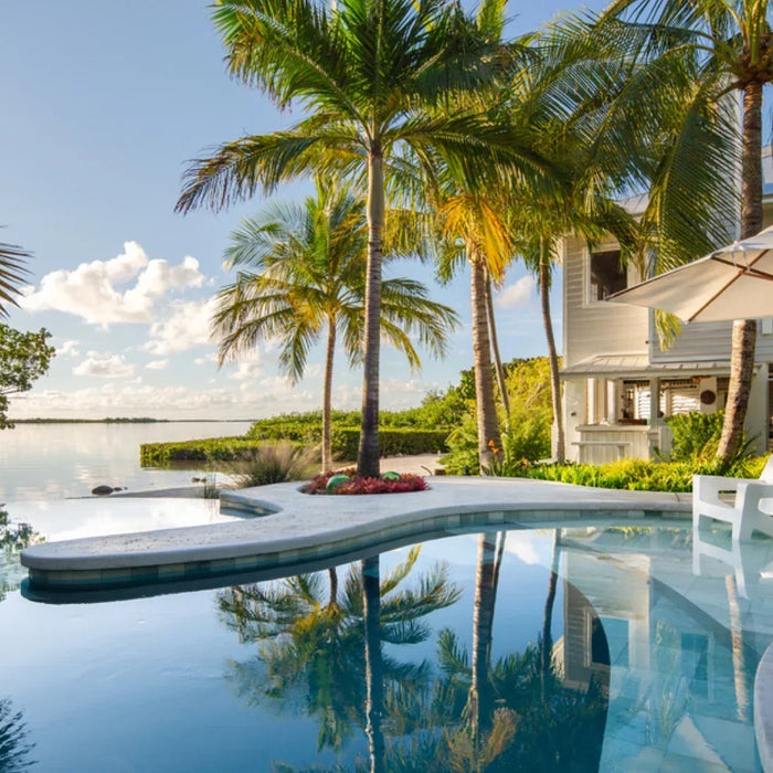 Modern backyard pool with two white in-pool lounge chairs and an umbrella overlooking tropical landscaping and open water.
