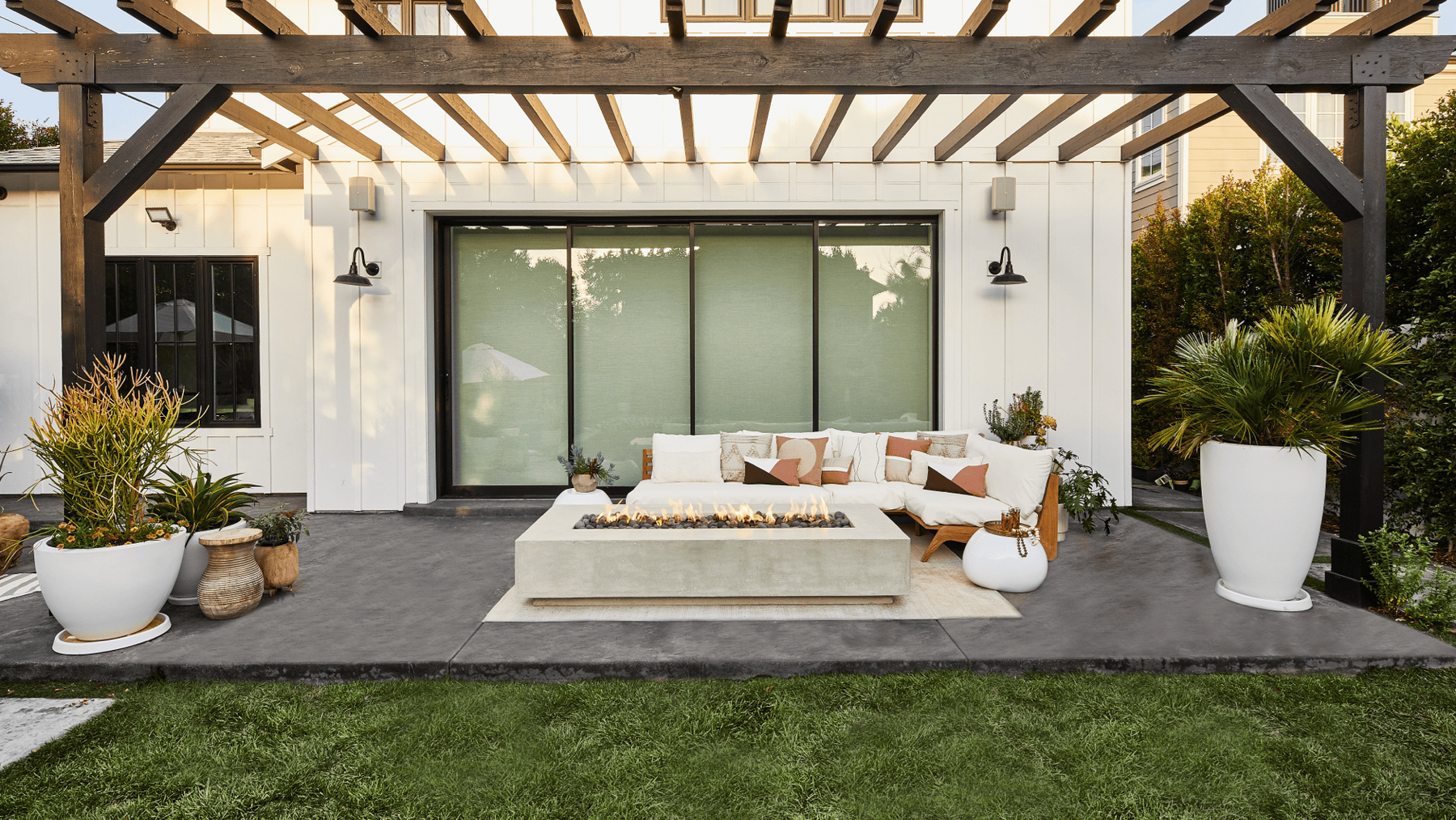 A modern outdoor patio with concrete fire pit, lounge sofa, and large potted plants all covered by a wooden pergola.