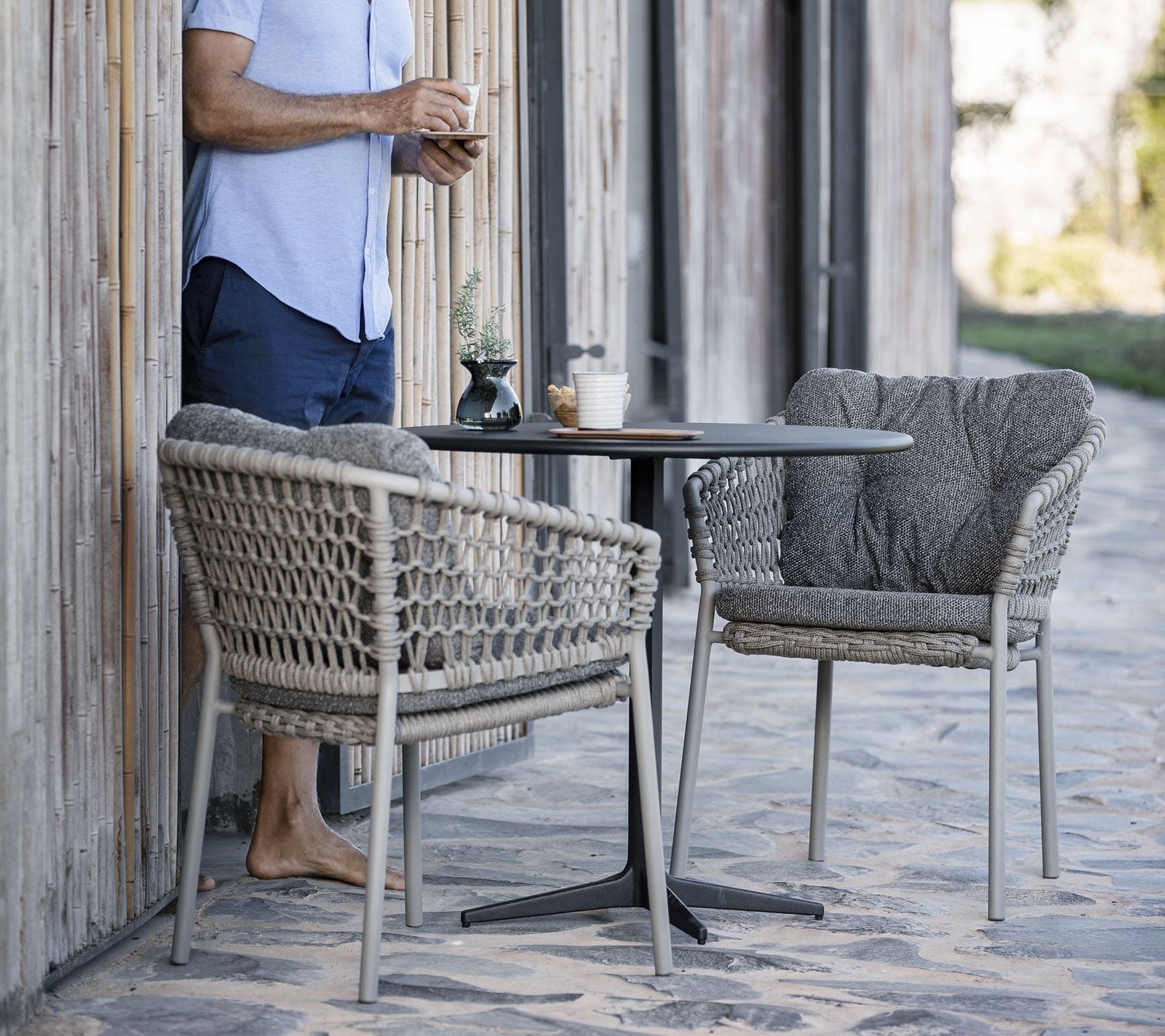 Boxhill's Drop Round Outdoor Cafe Table Lava Grey lifestyle image with 2 dining chairs and a man standing beside holding a cup