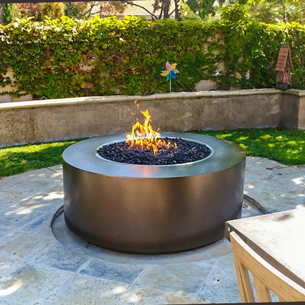 Dark brown round powdered coated fire pit with lava rocks and flames on top placed on a patio surrounded with green lawn