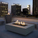 Gray outdoor concrete rectangular fire pit with flames set on a roof top patio with a view city skyline with tall buildings