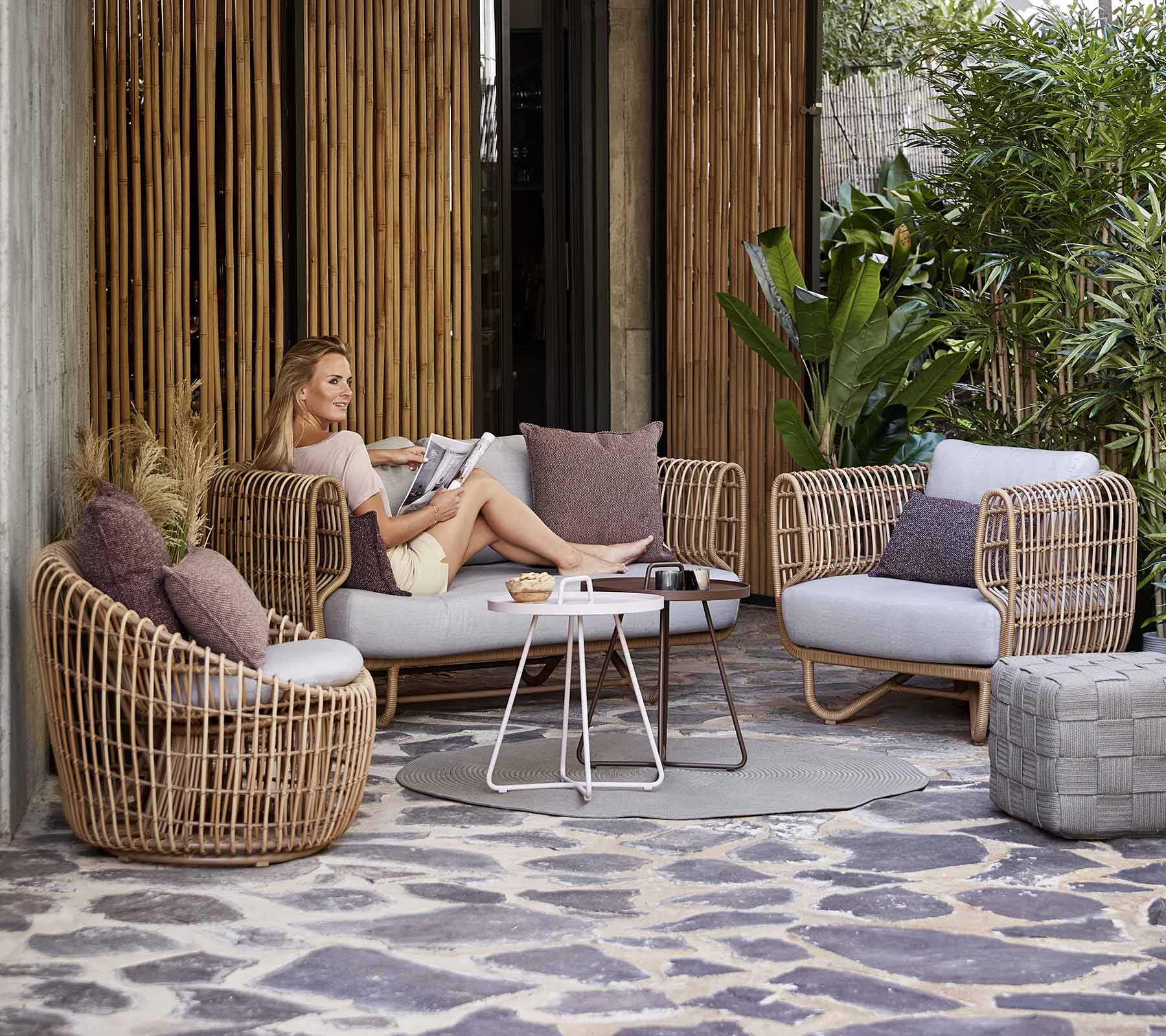 Boxhill's Nest Lounge Chair Lifestyle image with Nest 2-Seater Sofa, Nest Round Rattan Chair and a woman sitting and reading a magazine, at patio