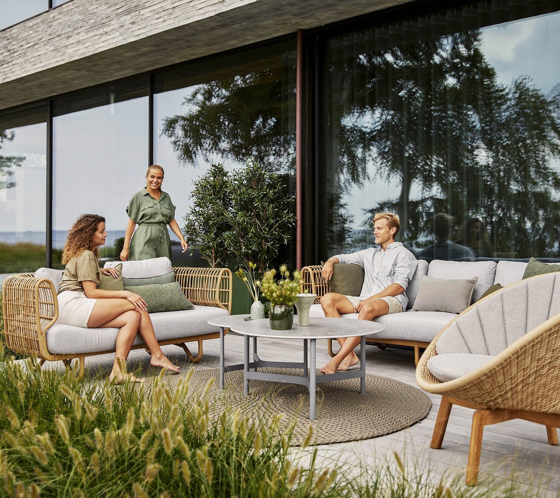Boxhill's Nest Sofa Outdoor 3 Seater lifestyle image at patio with man and a woman sitting down having a chat, and another woman walking at the side