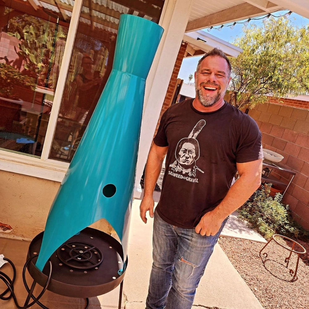 A man standing beside a tall, unique turquoise outdoor metal fire pit on a patio, set against large glass windows