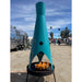 A tall modern outdoor metal fire pit with a unique turquoise design, placed on a sandy surface outdoors near a metallic bus