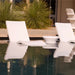 Two white tanning ledge chairs and small pool side table partially submerged in a pool, with tall green plants in the background.