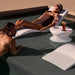 A woman in a brown swimsuit lounges on a white in-pool chaise lounger while a man leans on the pool edge near a table with drinks.