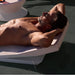 Man wearing sunglasses relaxing shirtless on a white in-pool chaise lounger, resting with his arms behind his head on a pool ledge.