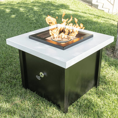 A black and white square outdoor metal powdered coated fire pit table with flames set on an outdoor grassy lawn area