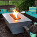 A grey tapered shaped concrete rectangular fire pit set on a patio surrounded by a wicker sofas and chair with green cushion