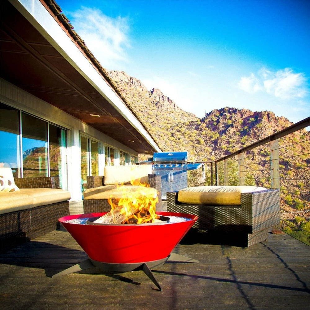 Red round tapered shaped metal fire pit with black steel legs with burning woods on it set on a patio with cushioned wicker sofa and chairs