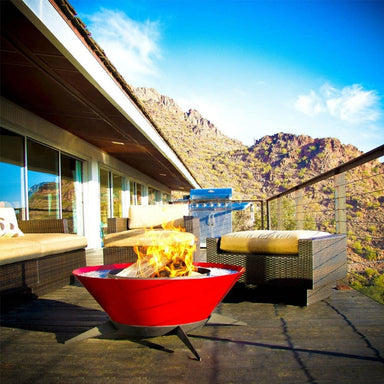 Red round tapered shaped metal fire pit with black steel legs with burning woods on it set on a patio with cushioned wicker sofa and chairs