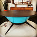 Light blue round tapered shaped metal fire pit with black steel legs with fire pit cover on a white circular platform in a room