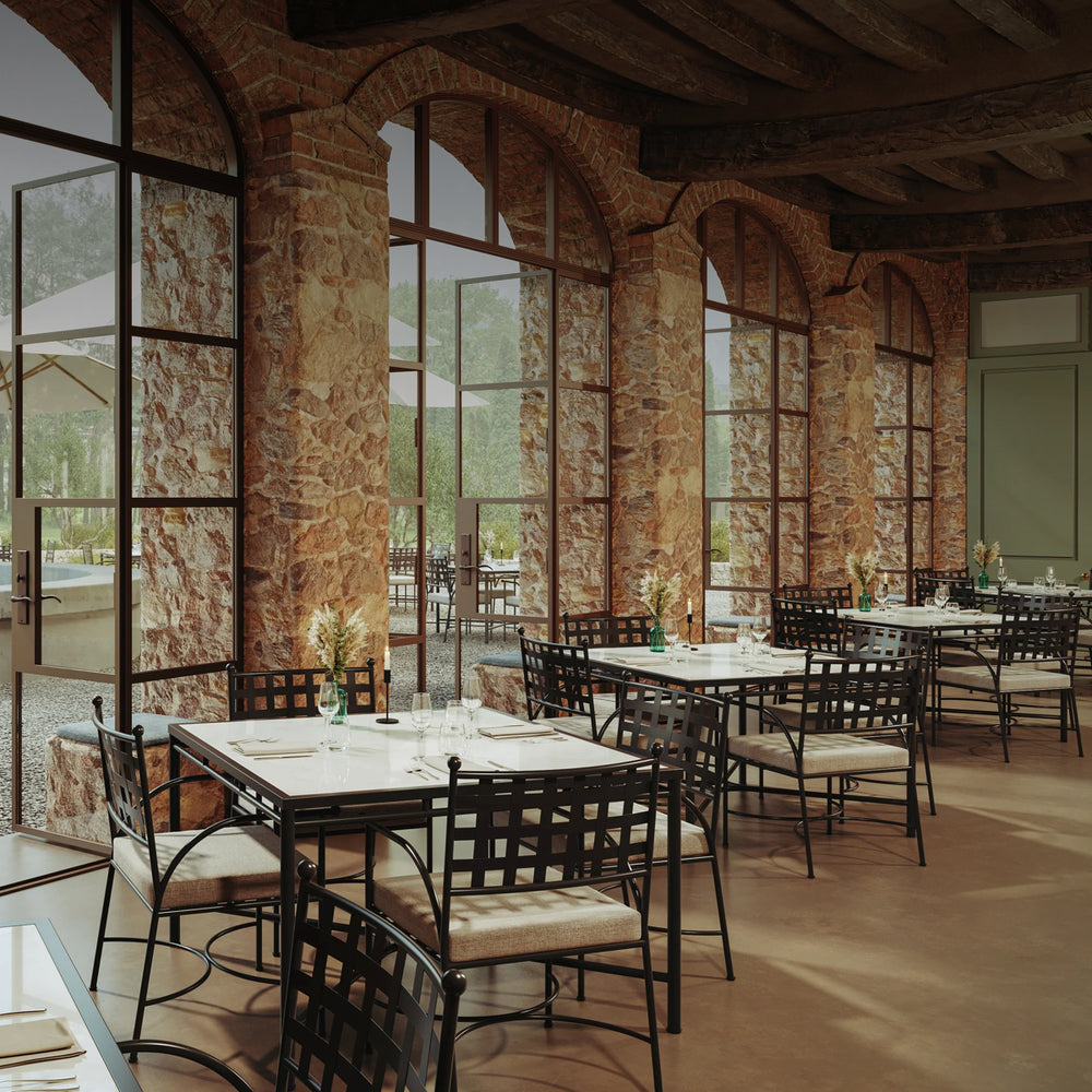 Cozy indoor/outdoor hospitality dining space with wrought iron chairs, stone walls, and large arched windows opening to an outdoor patio.