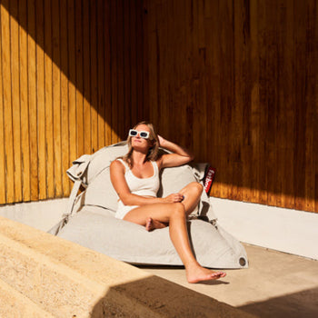 Woman relaxing on a modern outdoor in-pool bean bag chair in the sun, styled against a wood-paneled wall.
