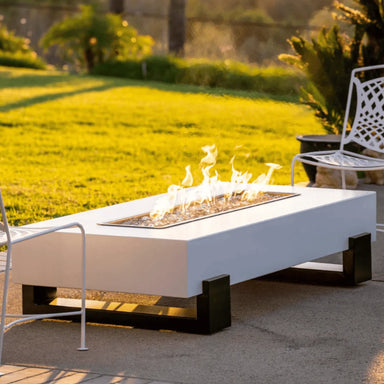 A white outdoor rectangular powder coated metal fire pit, and two white metal chairs set on a patio with a lush green lawn
