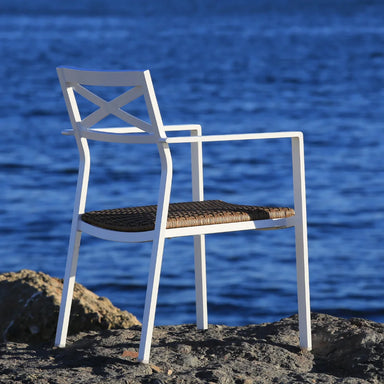 A white aluminum stackable outdoor dining armchair with wicker padded seat  placed on a rocky surface by the water