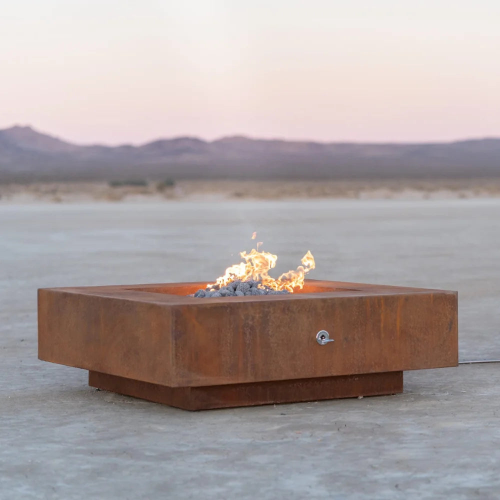 Corten steel square outdoor metal fire pits with lava rocks and flames placed on an outdoor area with a view of a distant mountain