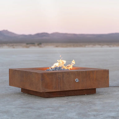 Corten steel square outdoor metal fire pits with lava rocks and flames placed on an outdoor area with a view of a distant mountain