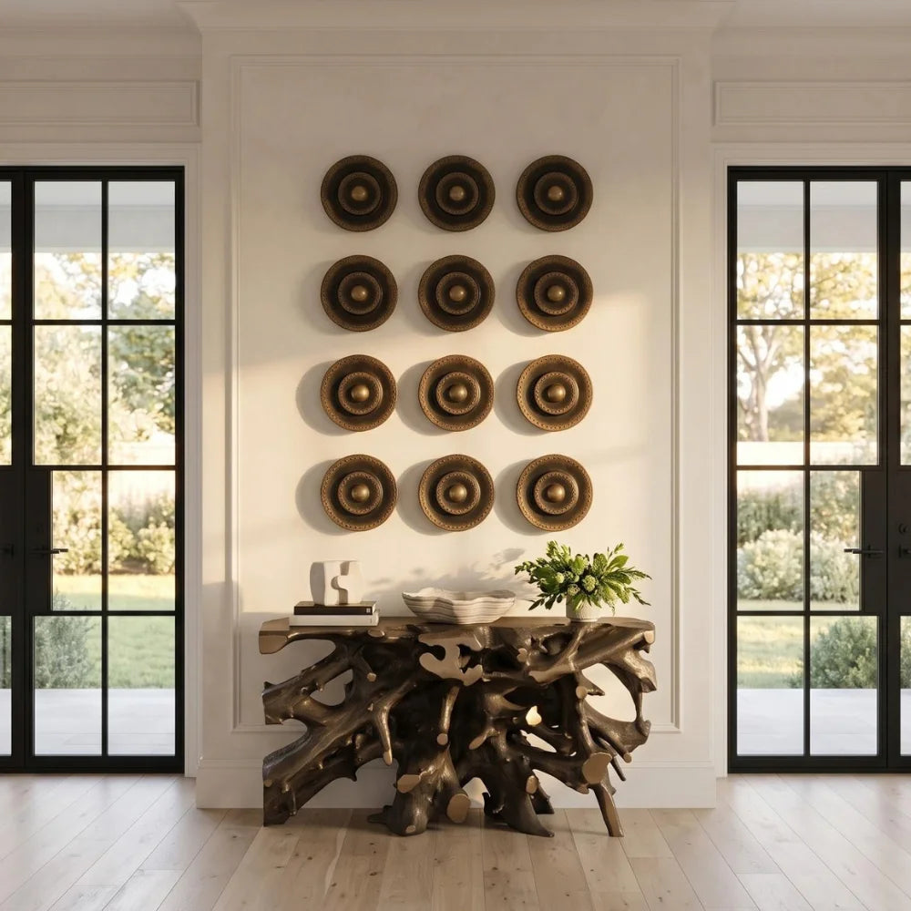 A styled interior scene showing a console table made from natural tree roots, decorated with a bowl and books, beneath a grid of twelve circular woven wall decor pieces. The room features light wood floors, white walls with molding, and black-framed glass doors looking out onto a green garden.