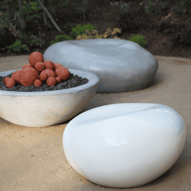 Light grey concrete pebble garden seat and white fiberglass pebble seat on a sandy surface with grey concrete fire bowl