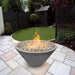 A gray tapered shaped round fire pit filled with lava rocks with flame situated on a tiled patio next to a green lawn area