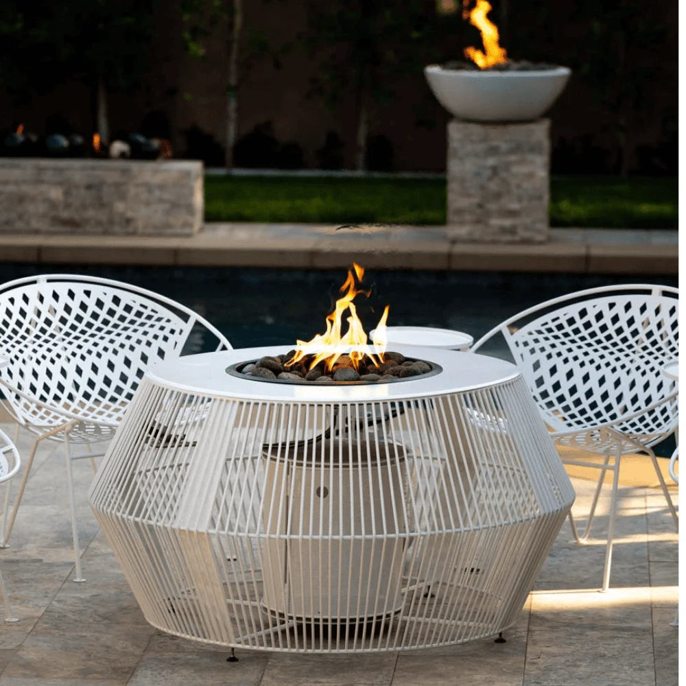 A white outdoor metal powdered coated fire pit table set on a patio beside the pool accompanied by two white metal chairs