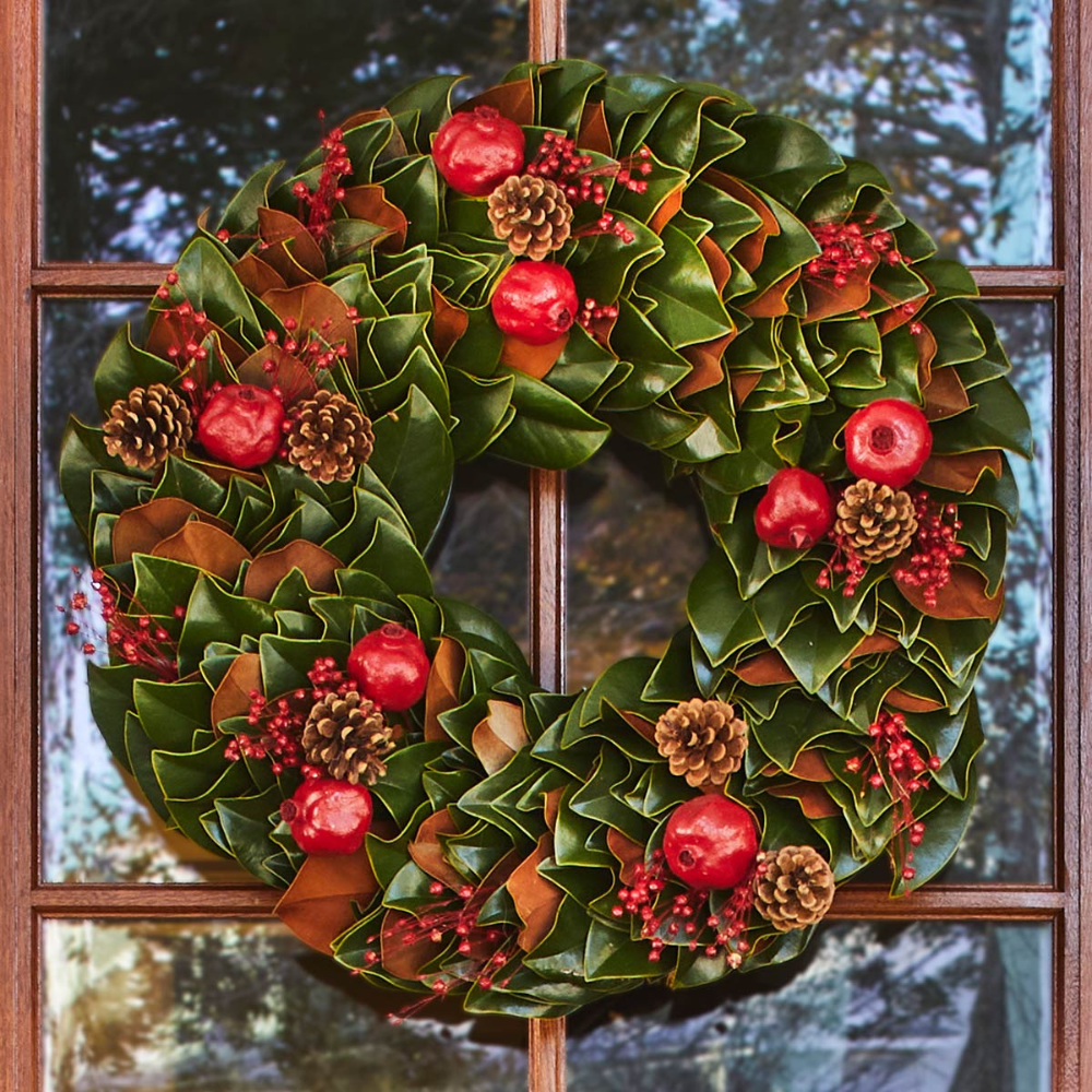 Christmas wreath with green leaves, red apples, and pinecones in front of a window.