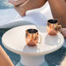 Two hammered copper Moscow mule mugs with gold-toned handles and stirrers, placed on a round white in-pool side table near a pool, with a person's legs and feet visible in the background.