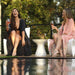 Two women sitting on white outdoor lounge chairs by a pool, holding wine glasses and wearing matching black and pink robes, with trees in the background.