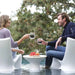 A man and a woman sitting in white outdoor lounge chairs at a pool, holding up clear glasses with dark beverages. A small round table between them holds fruit, with blurred trees in the background.