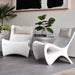 Two modern white sculptural in-pool lounge chairs with beige pillows, styled in a minimalist living room setting with a side table, potted plants, and a gray floor.