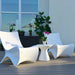 Two modern white sculptural lounge chairs with a matching small round side table on a wooden deck, with potted plants and a window in the background.