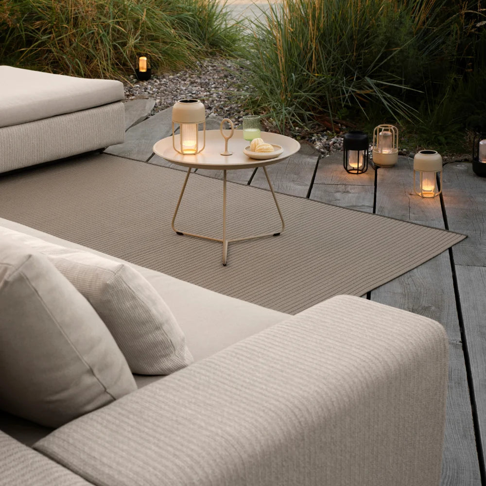 Outdoor patio with beige sectional sofa, brown outdoor rug, round tray table, and portable lamps surrounded by grasses