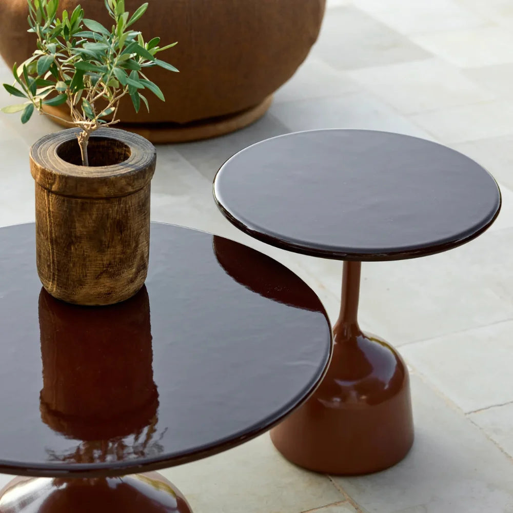 Two shiny dark brown round patio coffee tables on a tiled floor, one topped with a wooden pot containing plants