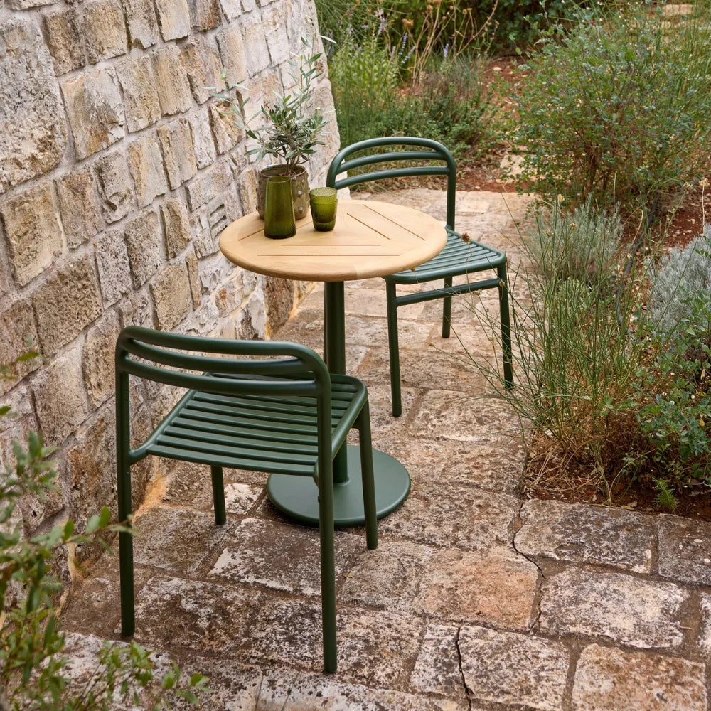 Round outdoor cafe table with wooden top and green metal base, paired with two green metal dining chairs on a patio against a concrete wall