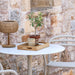 White aluminum patio cafe table with wooden tray, cup, potted plant, and wicker table lamp, set against a brick wall with a pair of white dining chairs