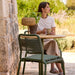 Outdoor patio with round outdoor cafe table with wooden top, paired with two green dining chairs, with a woman sitting on one chair holding a cup of coffee