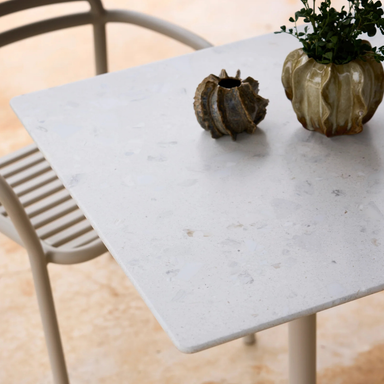 Close view of a white square outdoor cafe table with textured surface, two vases, and a chair on a patio