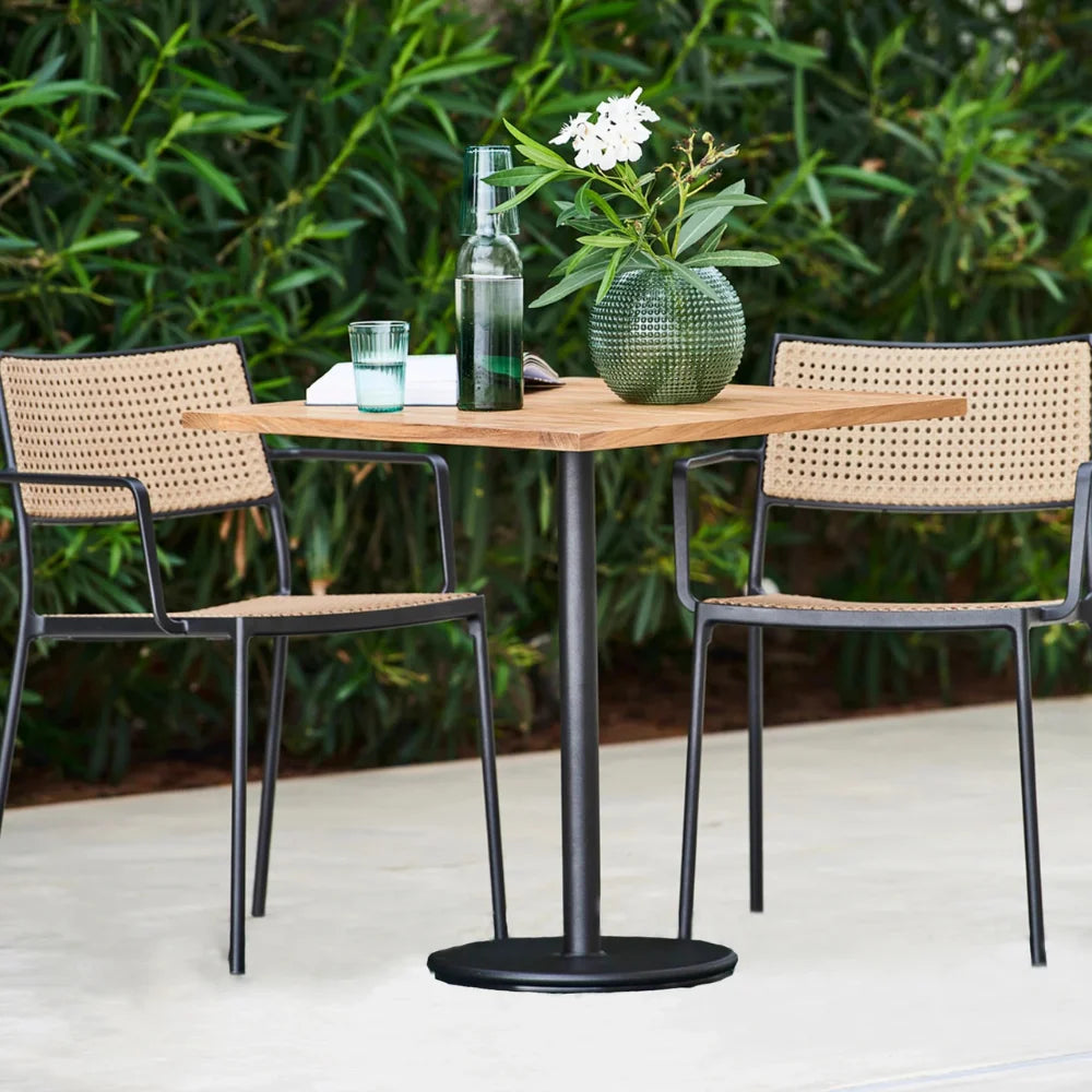 Square wooden-top outdoor cafe table with two outdoor chairs, a vase of flowers, a glass of water, surrounded by greenery on a patio