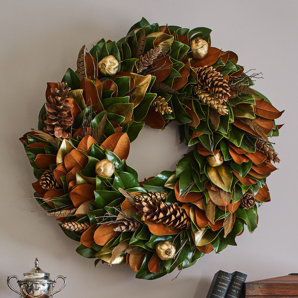 Decorative wreath with green leaves, pinecones, and gold balls on a plain wall.