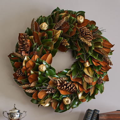 Decorative wreath with green leaves, pinecones, and gold balls on a plain wall.